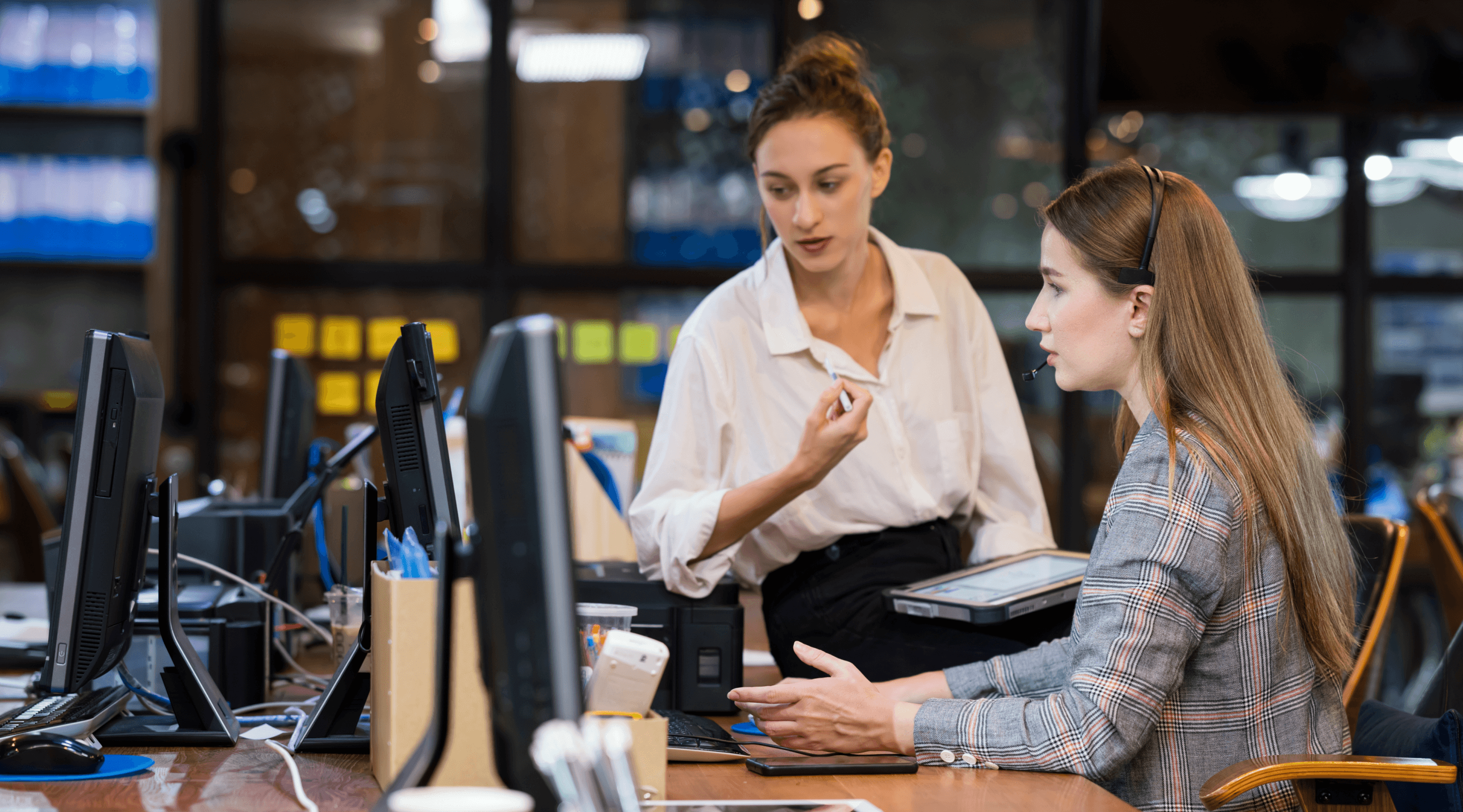 Two women talking over a desktop and looking at a computer screen.