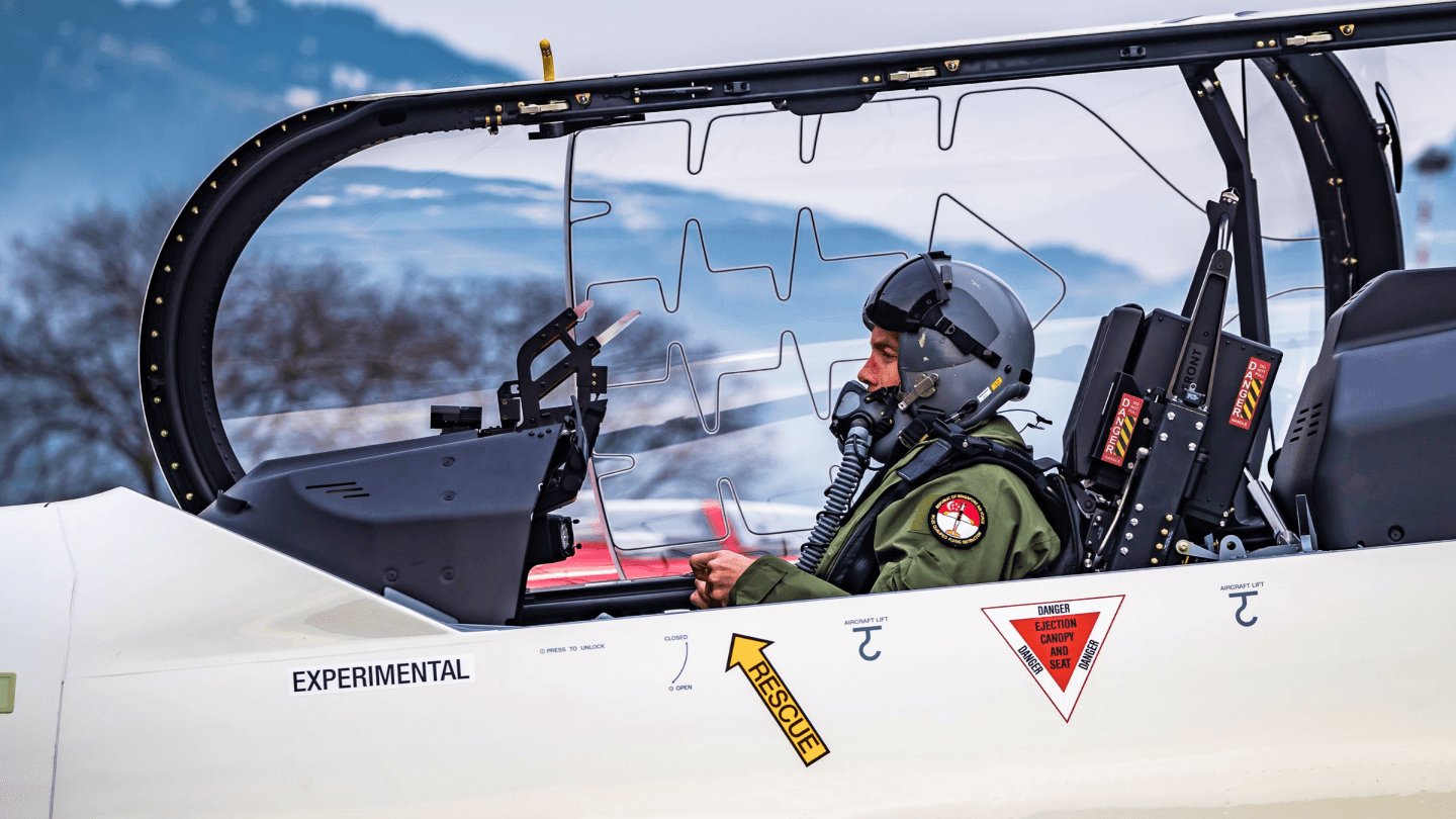 Un pilota con casco e maschera di ossigeno è seduto nella cabina di pilotaggio di un jet sperimentale bianco.
