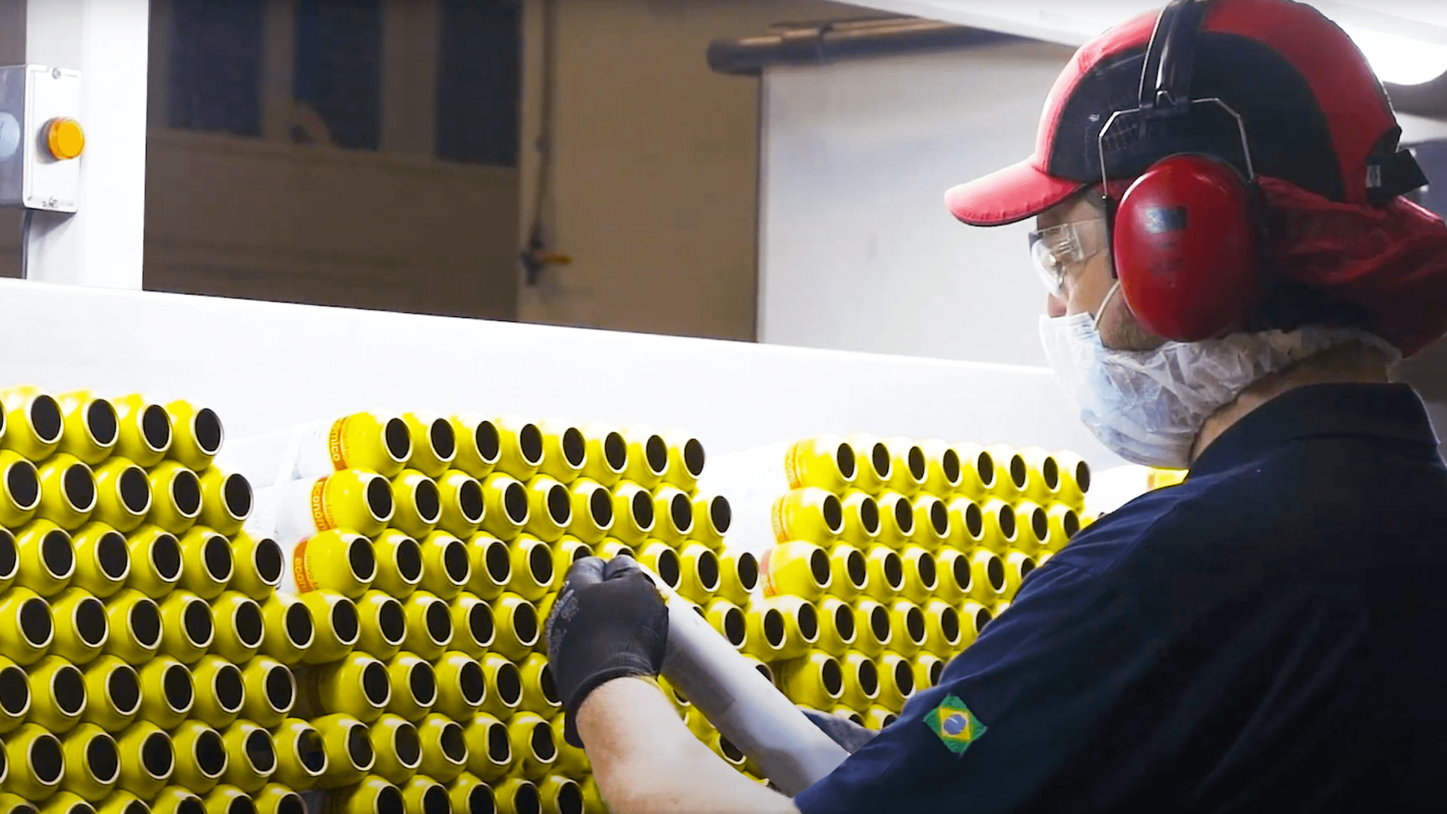 Un operaio con indosso indumenti protettivi maneggia file di lattine gialle in una fabbrica.
