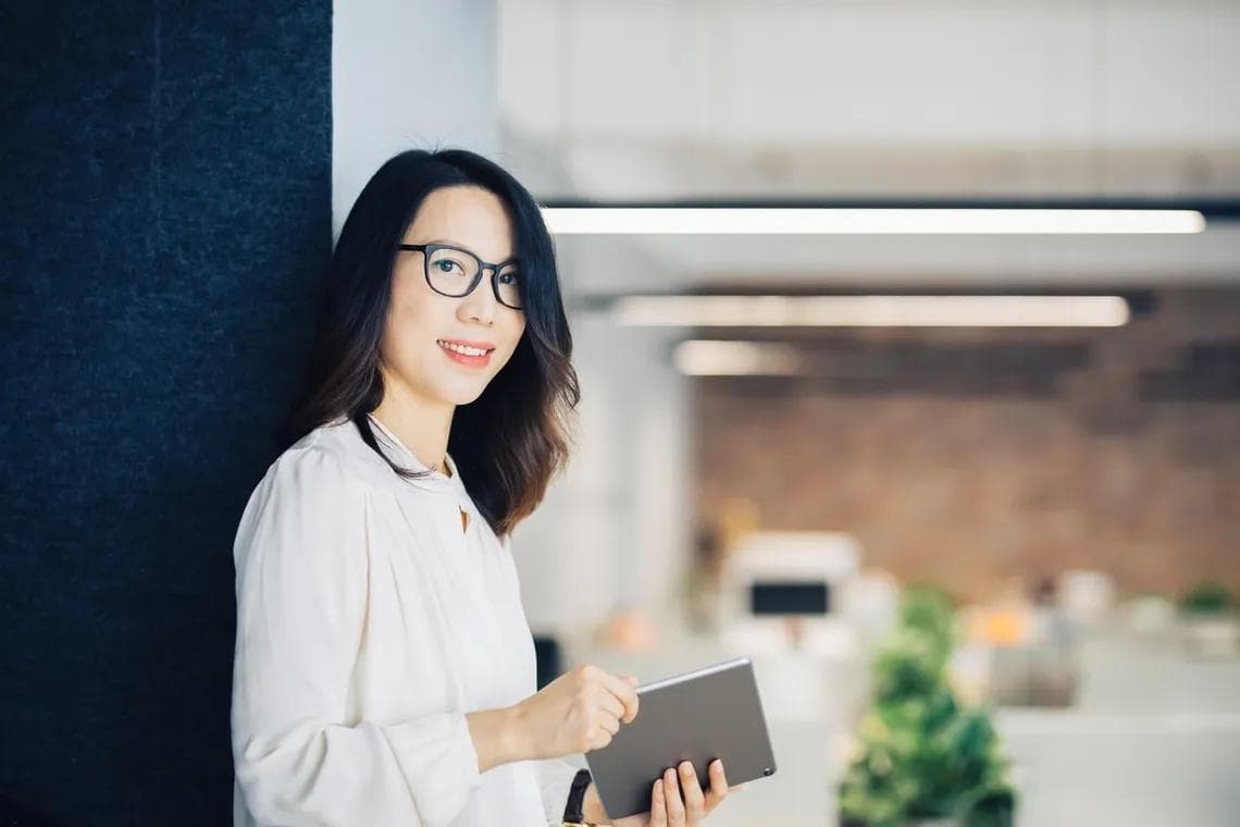 a woman wearing glasses is leaning against a wall while holding a tablet computer .