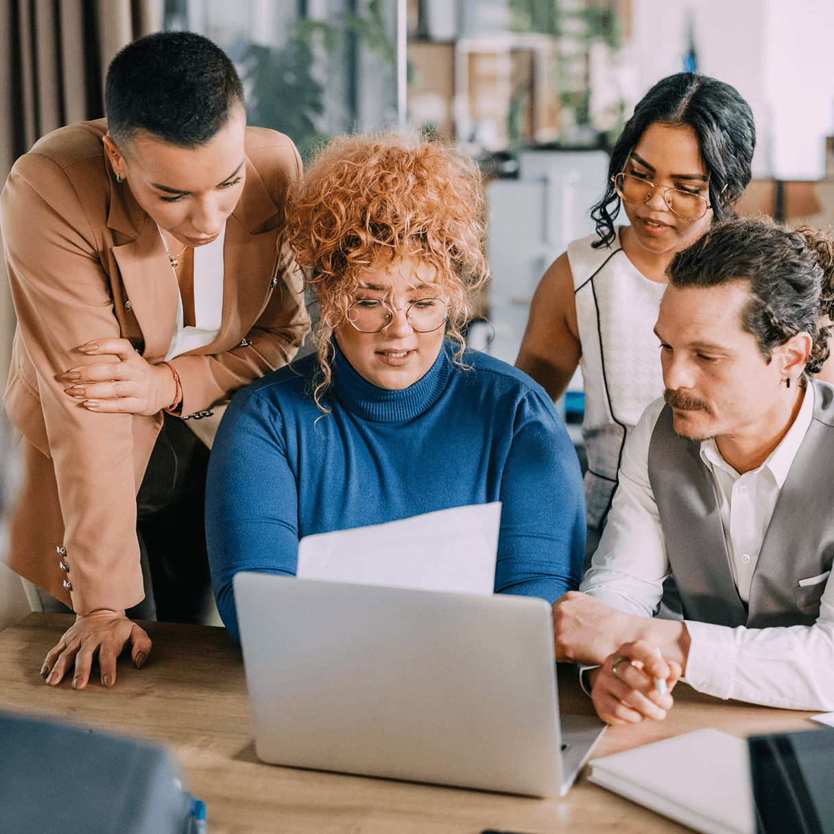 Four colleagues gather closely around a desk, looking intently at a laptop screen and papers during a collaborative meeting.
