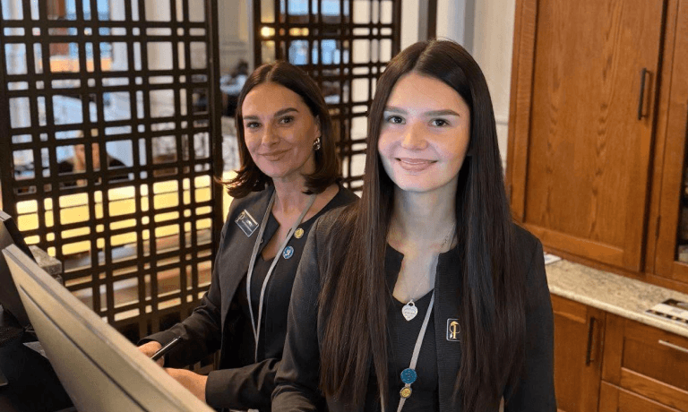 Two smiling women in black blazers and lanyards stand in a reception area.