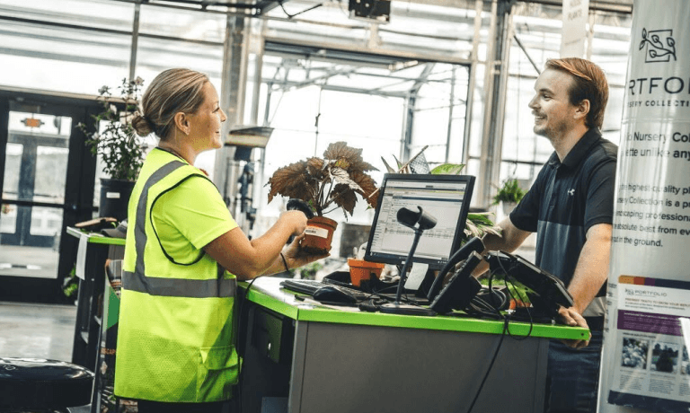A woman in a safety vest scans a potted plant while a man smiles at her from behind the counter in a plant nursery.