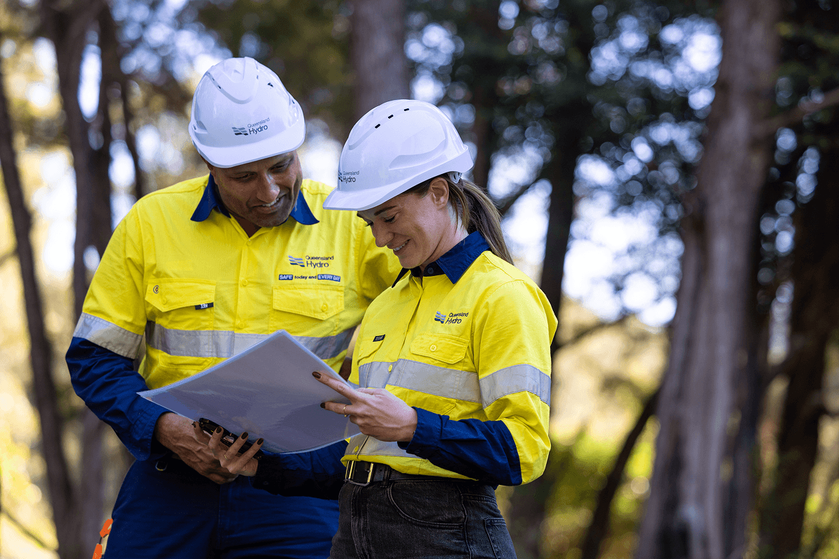 Two Queensland Hydro workers in hard hats and hi-vis shirts reviewing documents outdoors.