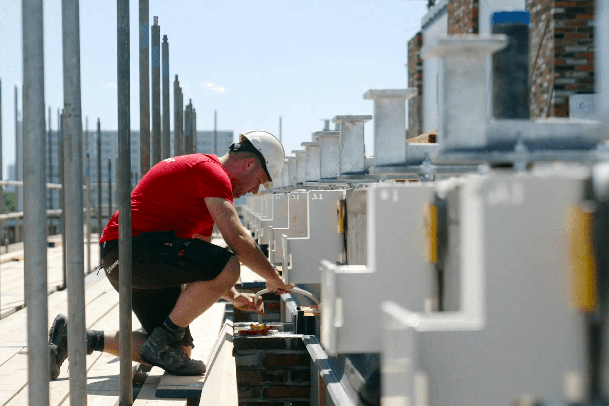 A construction worker in a hard hat and red shirt kneels on scaffolding, installing components.