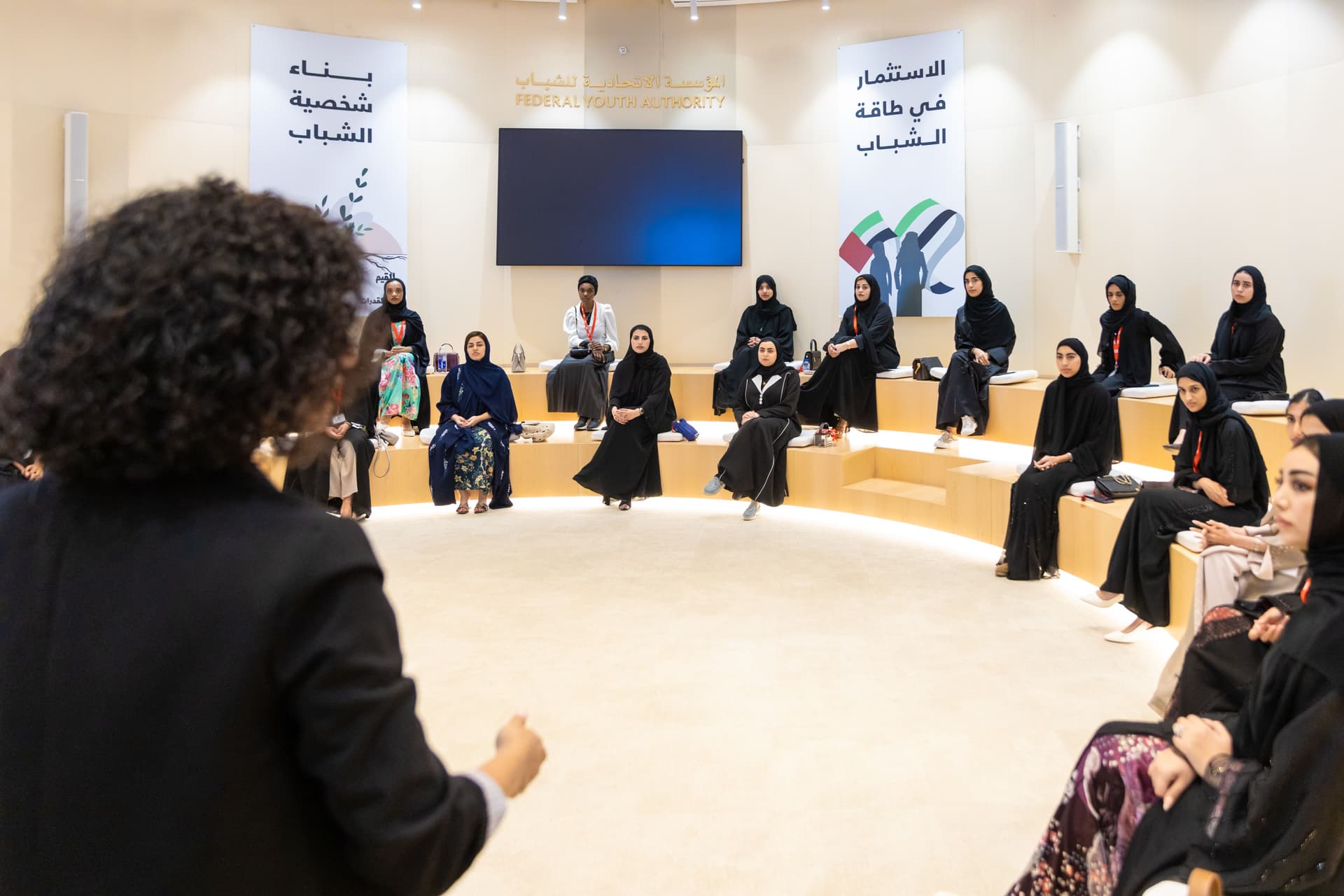 A woman speaks to a group of young women, most wearing abayas and hijabs, seated in a semi-circle in a modern room with "Federal Youth Authority" banners.