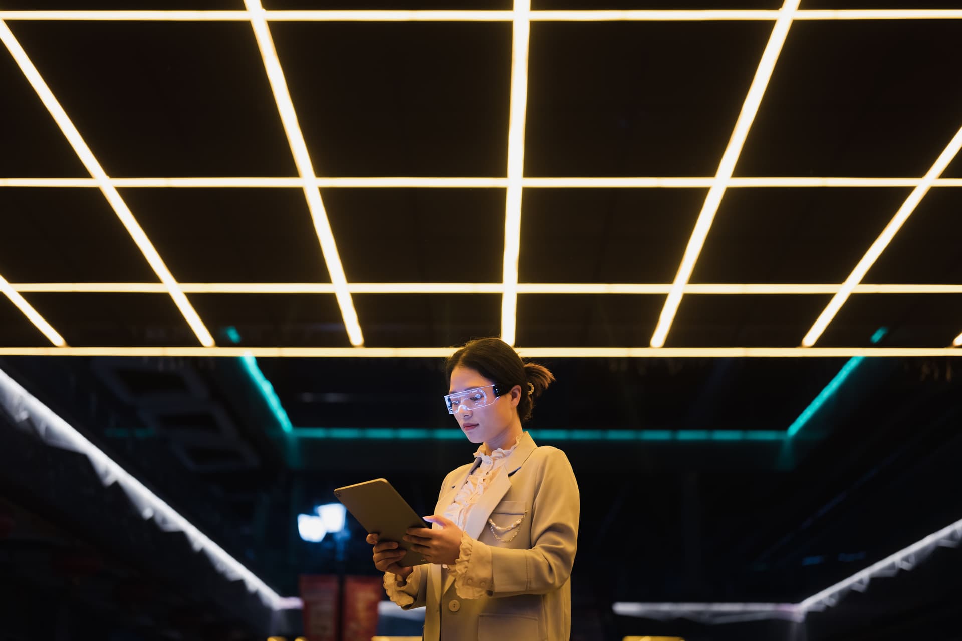 Woman with glowing AR glasses holding a tablet under a grid of neon ceiling lights.