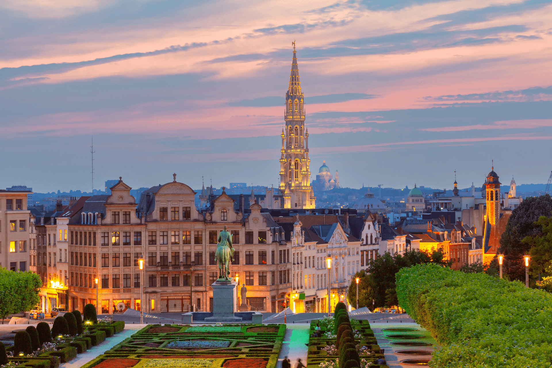 Brussels cityscape at dusk, featuring the illuminated Town Hall spire and a formal garden in the foreground.