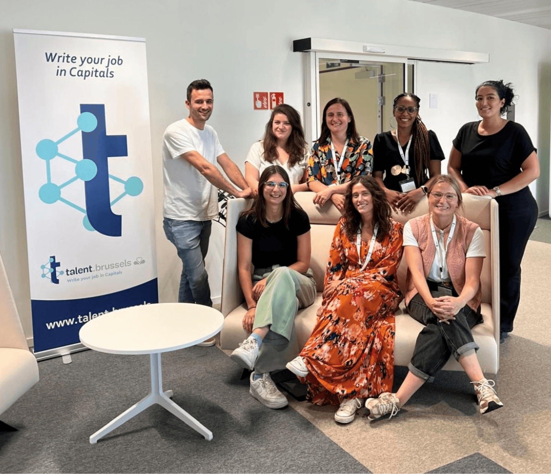 Eight smiling people, one man and seven women, pose in an office with a "talent.brussels" banner.