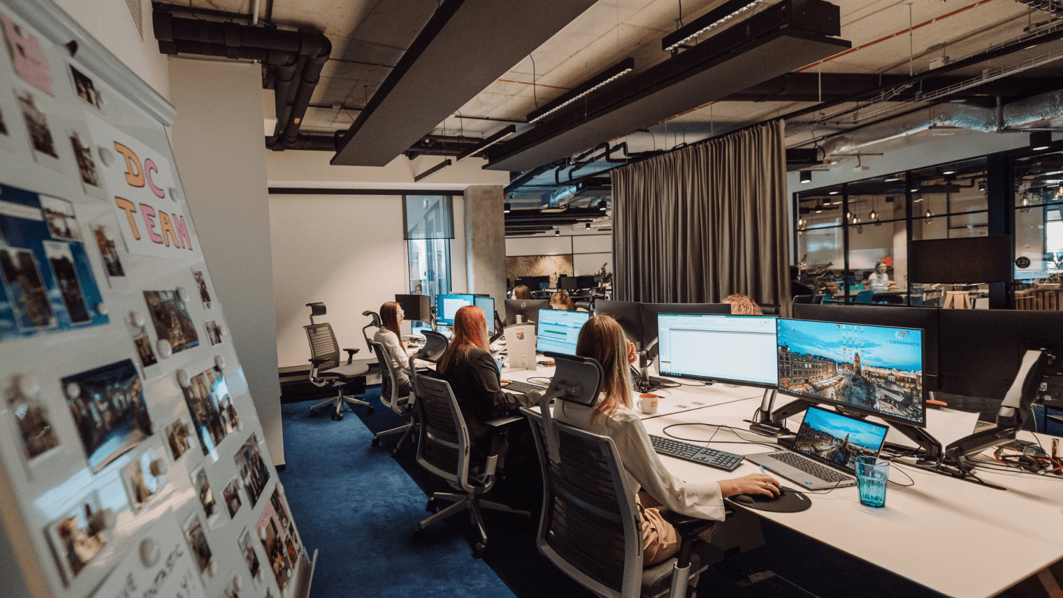 Image of people working in an office. The words "DC team" appear on a wall with pictures behind them.