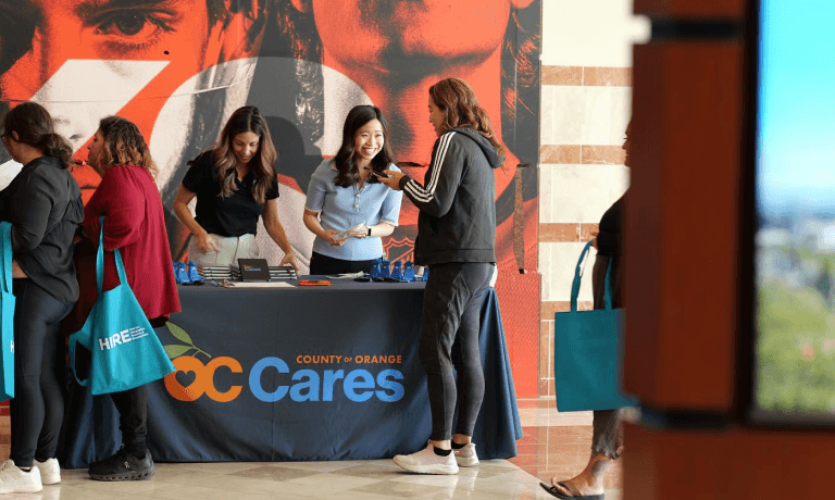 Frauen unterhalten sich an einem Stand für die Veranstaltung „County of Orange OC Cares“, wobei einige „HIRE“-Einkaufstaschen tragen.