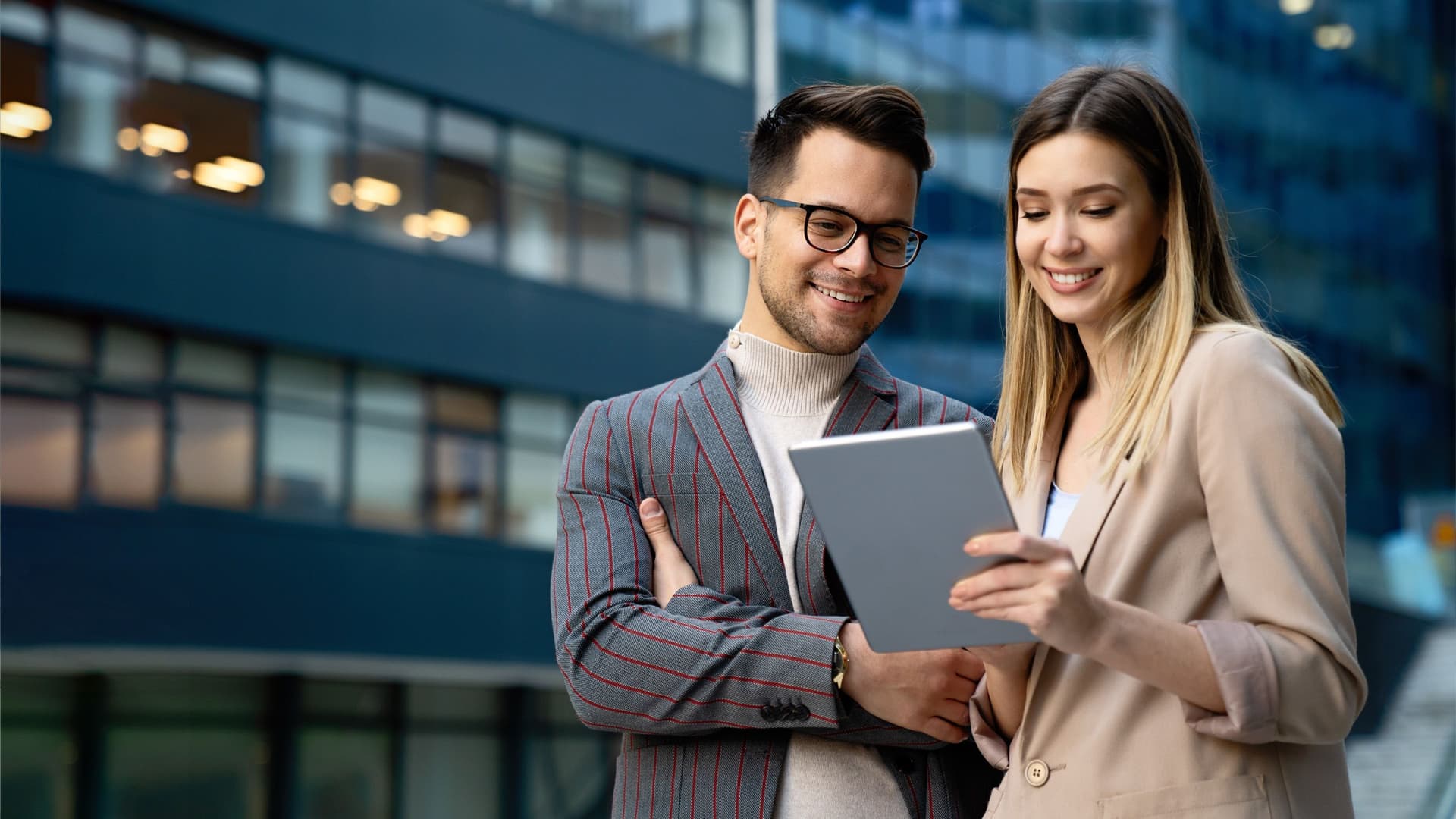 Two smiling business people looking at a tablet in front of a modern building.