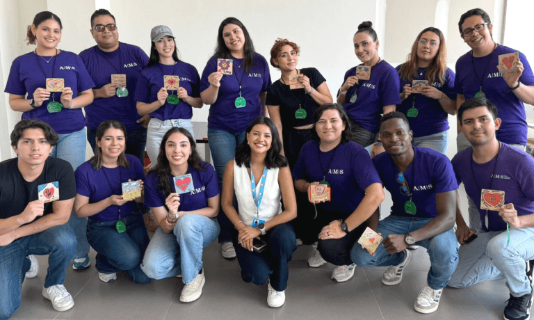 Un grupo diverso de personas sonrientes, muchas con camisetas moradas de AMS, que sostienen unas pequeñas tarjetas decoradas.