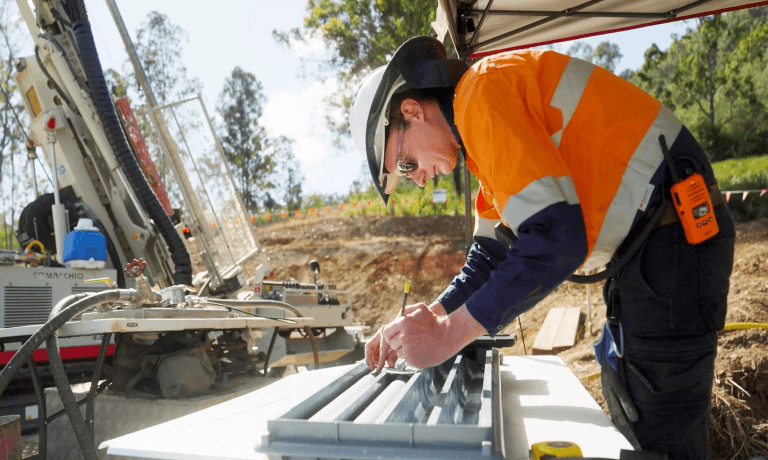 A person in a hard hat and high-vis jacket examines geological core samples, with a drilling rig behind them.