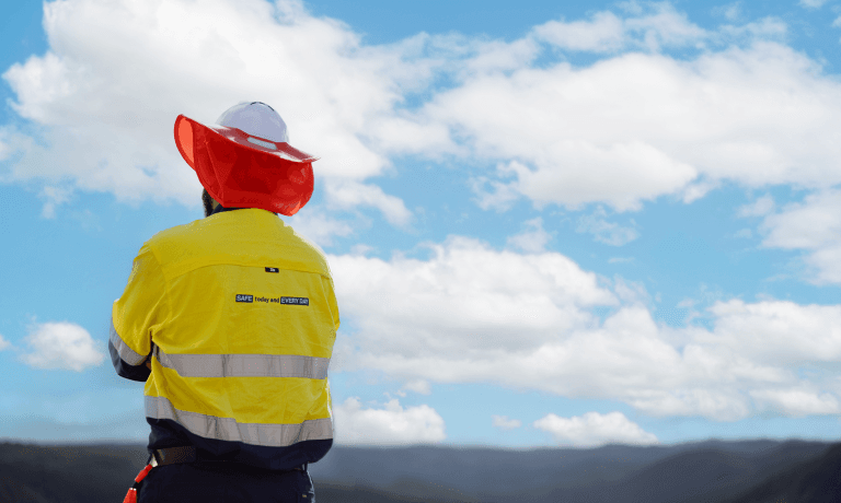 A person in a yellow high-visibility jacket and hard hat looks out at a landscape under a blue, cloudy sky.