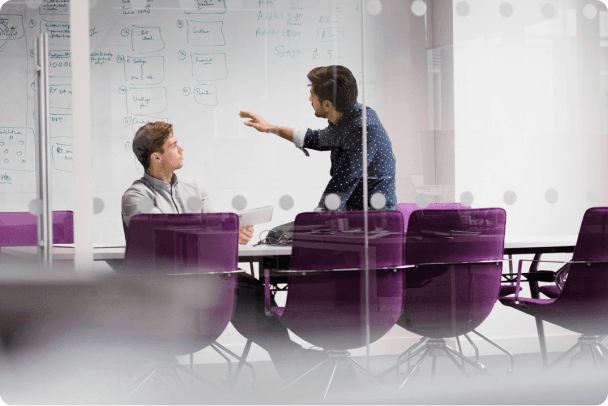 Two men discussing in a modern meeting room with a whiteboard and purple chairs.