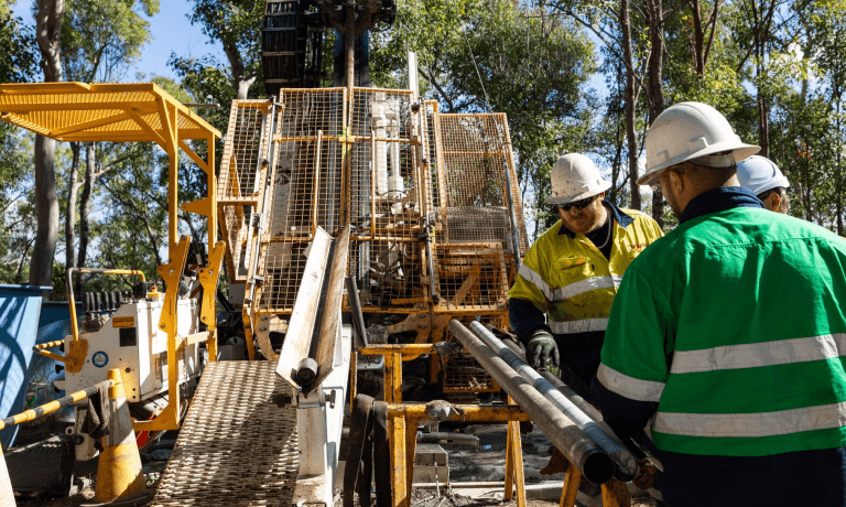 Three workers in hard hats handle drill pipes next to a drilling rig.