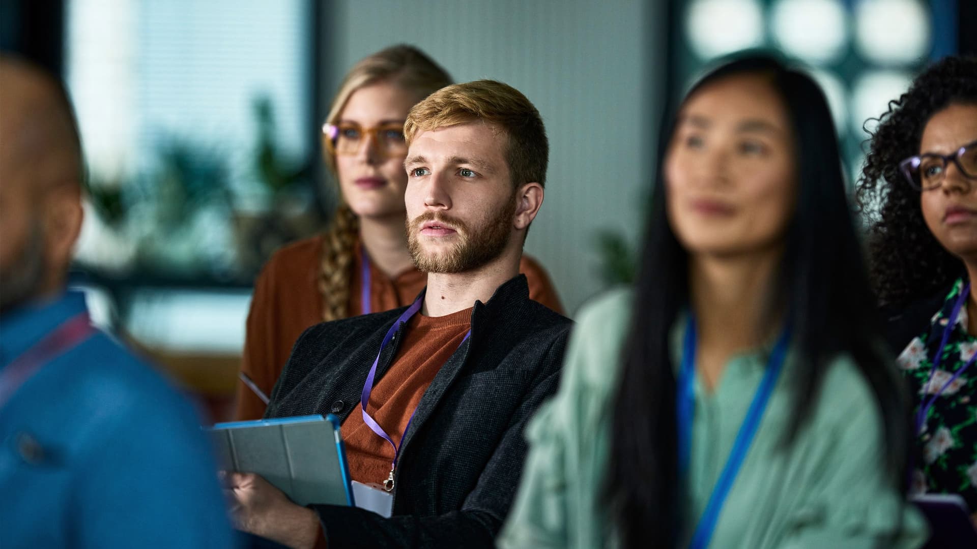 Diverse conference attendees, including a man holding a tablet, listen attentively.