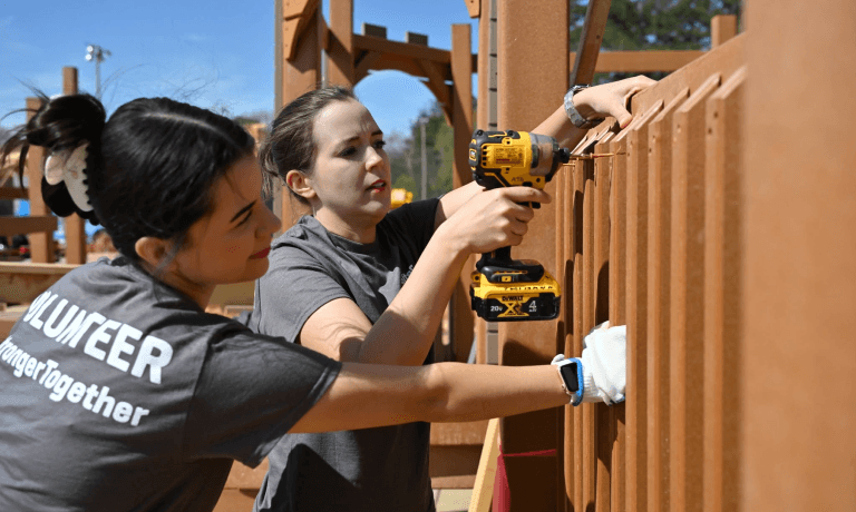 Two women, one wearing a "VOLUNTEER" shirt, use a drill to assemble a wooden structure outdoors.