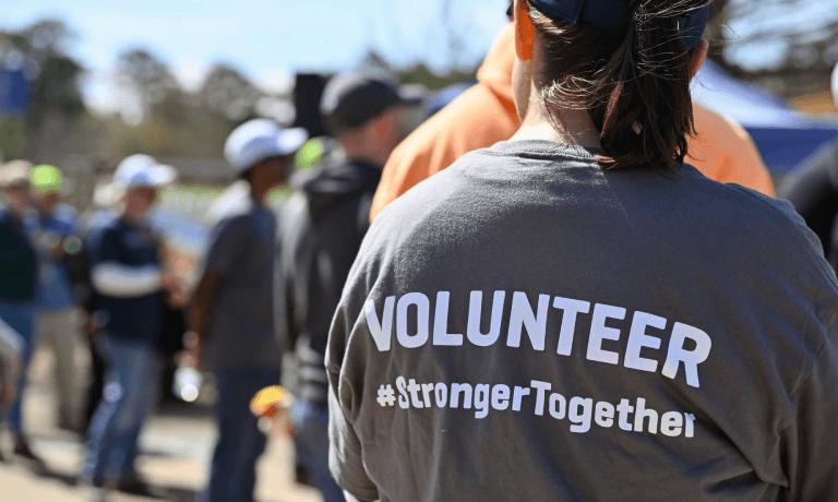 Back of a person wearing a grey t-shirt that says "VOLUNTEER #StrongerTogether" amidst a blurred crowd.