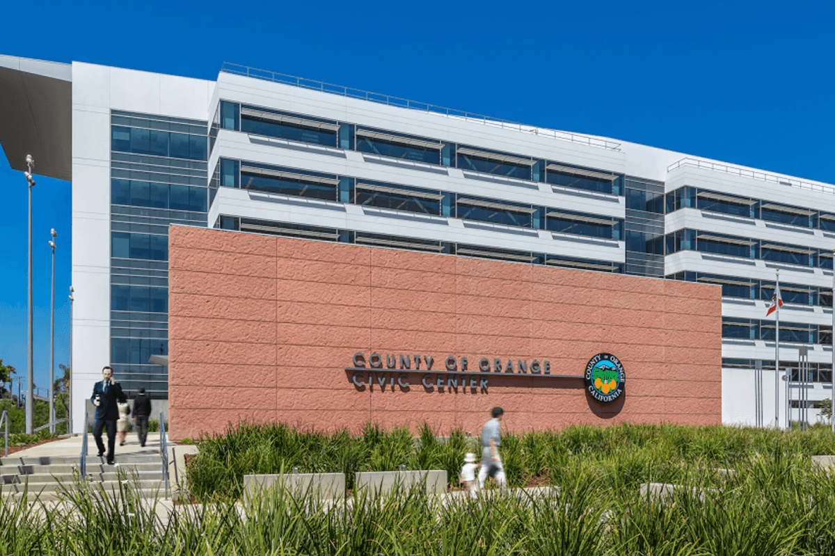 Ein modernes, weißes Gebäude, das County of Orange Civic Center, mit einer großen rot-braunen Beschriftungswand davor, umgeben von Grünanlagen und verschwommenen vorbeigehenden Personen.